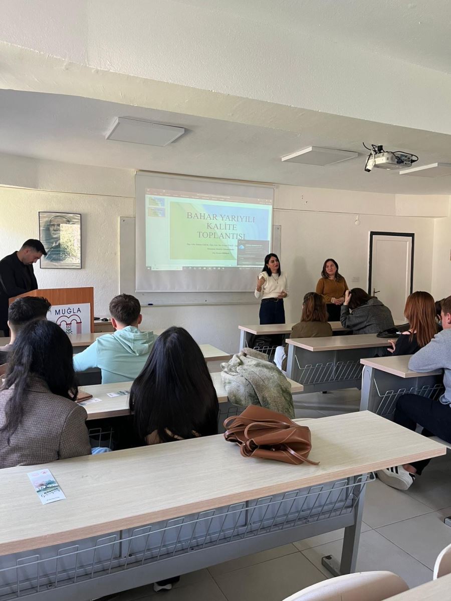 In a university classroom, a quality meeting is being held. Two instructors are standing at the front of the class while students are seated at desks listening attentively. A presentation titled “Spring Semester Quality Meeting” is projected on the screen. At the front of the classroom, a podium with the logo of Muğla Sıtkı Koçman University is visible. The instructors are addressing the students and leading the meeting.